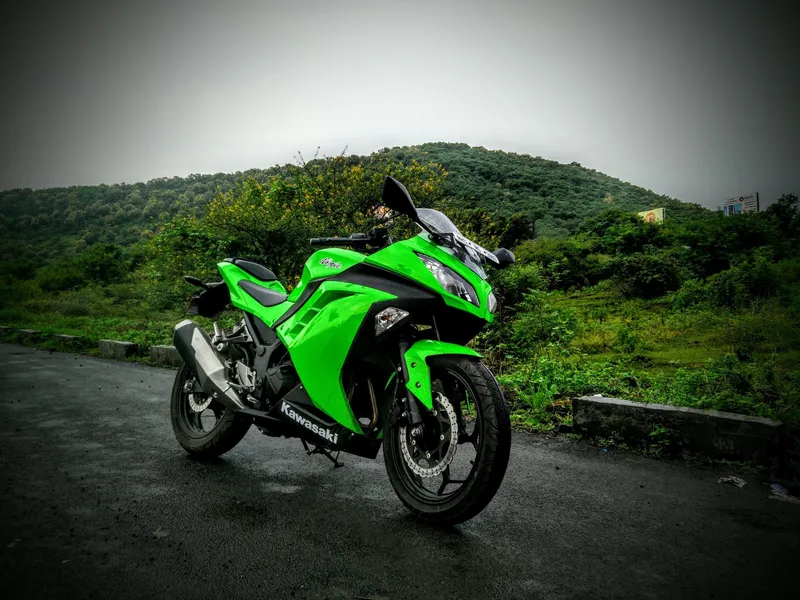 The green Kawasaki Ninja 300 parked on a wet hill road, monsoon greenery and low cloud behind it