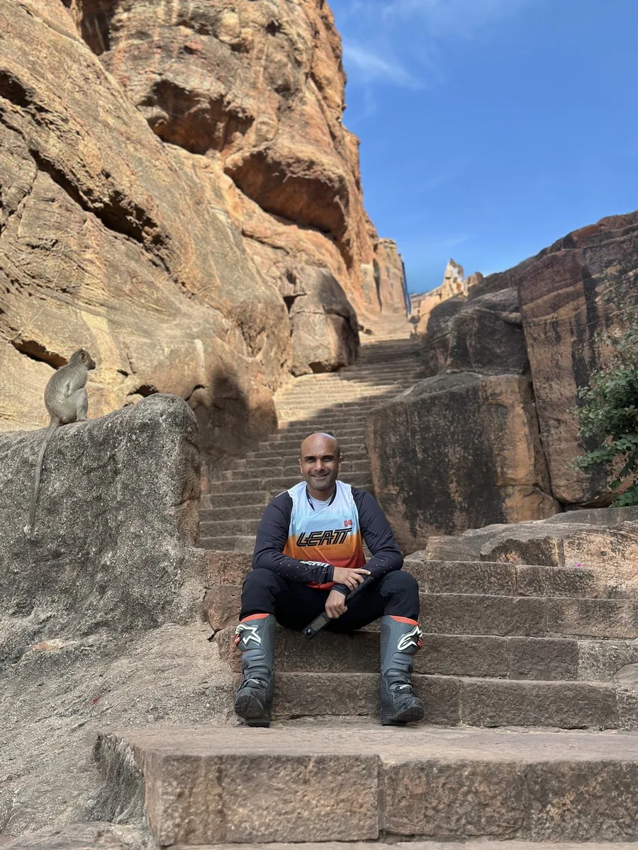 Leo sitting on the temple steps at Badami in riding gear, a monkey watching from the rock beside him